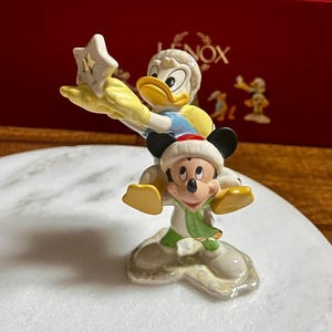 Może przedstawiać: Ceramiczna figurka przedstawiająca Kaczora Donalda trzymającego gwiazdę i Myszka Miki w czapce i szaliku Świętego Mikołaja. Figurka stoi na białej powierzchni, z czerwoną skrzynką w tle.