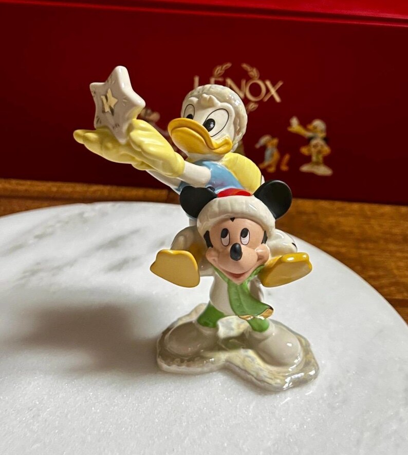 Może przedstawiać: Ceramiczna figurka przedstawiająca Kaczora Donalda trzymającego gwiazdę i Myszka Miki w czapce i szaliku Świętego Mikołaja. Figurka stoi na białej powierzchni, z czerwoną skrzynką w tle.