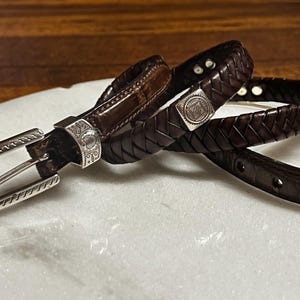 K&ouml;nnte beinhalten: Zwei braune Lederg&uuml;rtel mit silbernen Schnallen und Verzierungen. Ein G&uuml;rtel ist geflochten, der andere hat eine glatte Oberfl&auml;che. Die Schnallen und Verzierungen weisen aufwendige Designs auf. Die G&uuml;rtel werden auf einer wei&szlig;en Oberfl&auml;che pr&auml;sentiert.