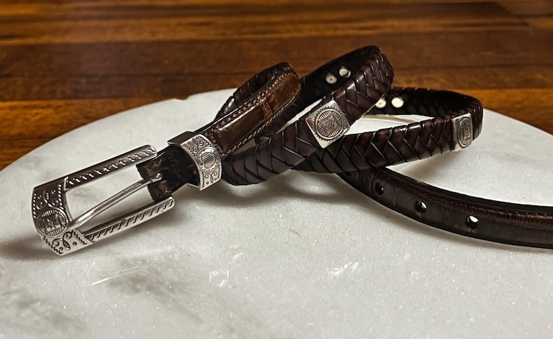 K&ouml;nnte beinhalten: Zwei braune Lederg&uuml;rtel mit silbernen Schnallen und Verzierungen. Ein G&uuml;rtel ist geflochten, der andere hat eine glatte Oberfl&auml;che. Die Schnallen und Verzierungen weisen aufwendige Designs auf. Die G&uuml;rtel werden auf einer wei&szlig;en Oberfl&auml;che pr&auml;sentiert.