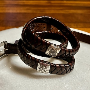 K&ouml;nnte beinhalten: Nahaufnahme von zwei dunkelbraunen Lederg&uuml;rteln mit geflochtenem Design. Jeder G&uuml;rtel hat eine silberfarbene quadratische Schnalle mit einem eingravierten Blumenmuster. Die G&uuml;rtel sind auf einer wei&szlig;en Oberfl&auml;che aufgerollt.