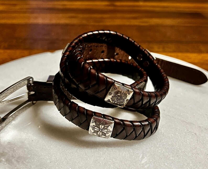 K&ouml;nnte beinhalten: Nahaufnahme von zwei dunkelbraunen Lederg&uuml;rteln mit geflochtenem Design. Jeder G&uuml;rtel hat eine silberfarbene quadratische Schnalle mit einem eingravierten Blumenmuster. Die G&uuml;rtel sind auf einer wei&szlig;en Oberfl&auml;che aufgerollt.