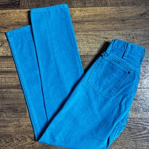 K&ouml;nnte beinhalten: Helle t&uuml;rkisfarbene Jeans mit klassischem F&uuml;nf-Taschen-Design. Die Jeans haben einen leuchtenden Blauton und scheinen ungetragen zu sein. Die Jeans liegen flach auf einer Holzoberfl&auml;che.
