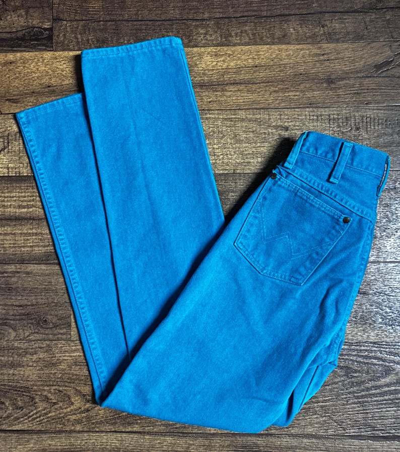 K&ouml;nnte beinhalten: Helle t&uuml;rkisfarbene Jeans mit klassischem F&uuml;nf-Taschen-Design. Die Jeans haben einen leuchtenden Blauton und scheinen ungetragen zu sein. Die Jeans liegen flach auf einer Holzoberfl&auml;che.