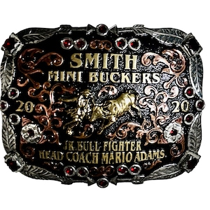 May include: A rectangular belt buckle with a black background, silver leaf-patterned border, and red rhinestones. It features gold-toned text reading SMITH MINI BUCKERS, 2020, JR. BULL FIGHTER, and HEAD COACH MARIO ADAMS, with a central bull rider figure.