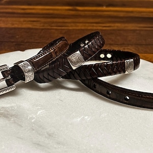 K&ouml;nnte beinhalten: Ein dunkelbrauner Lederg&uuml;rtel mit silberner Schnalle und Akzenten. Der G&uuml;rtel hat ein geflochtenes Design und eine rechteckige Silberschnalle mit dekorativen Details. Der G&uuml;rtel wird auf einer wei&szlig;en Oberfl&auml;che pr&auml;sentiert.