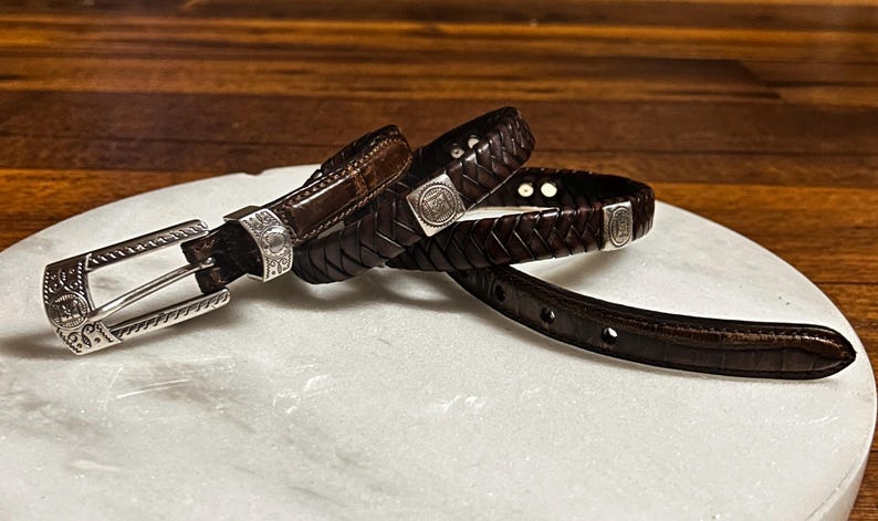 K&ouml;nnte beinhalten: Ein dunkelbrauner Lederg&uuml;rtel mit silberner Schnalle und Akzenten. Der G&uuml;rtel hat ein geflochtenes Design und eine rechteckige Silberschnalle mit dekorativen Details. Der G&uuml;rtel wird auf einer wei&szlig;en Oberfl&auml;che pr&auml;sentiert.