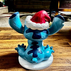 May include: A blue figurine of a cartoon character with large ears and a Santa hat. The figurine is sitting on a white marble base. The character has dark blue markings and is in a seated position with arms outstretched.