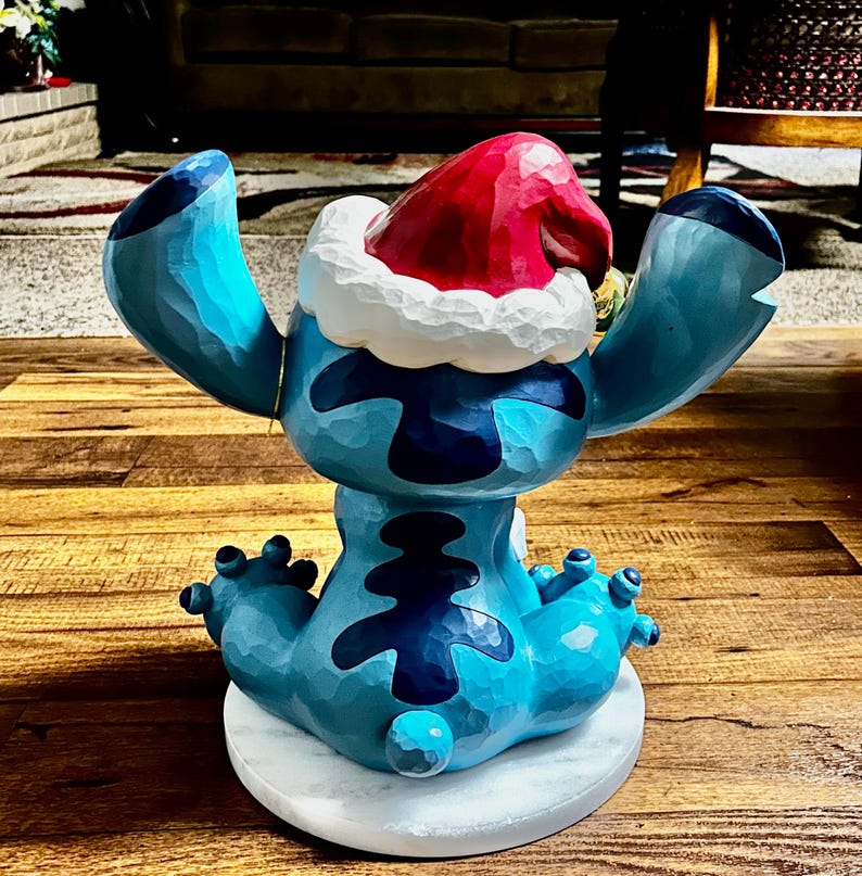 May include: A blue figurine of a cartoon character with large ears and a Santa hat. The figurine is sitting on a white marble base. The character has dark blue markings and is in a seated position with arms outstretched.