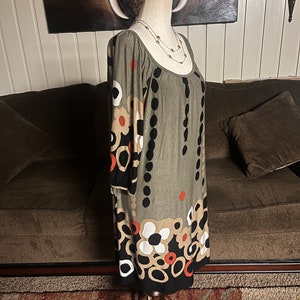 May include: A green, black, white, and orange patterned dress with a scoop neck and 3/4 sleeves. The dress is on a mannequin and is displayed against a brown couch.