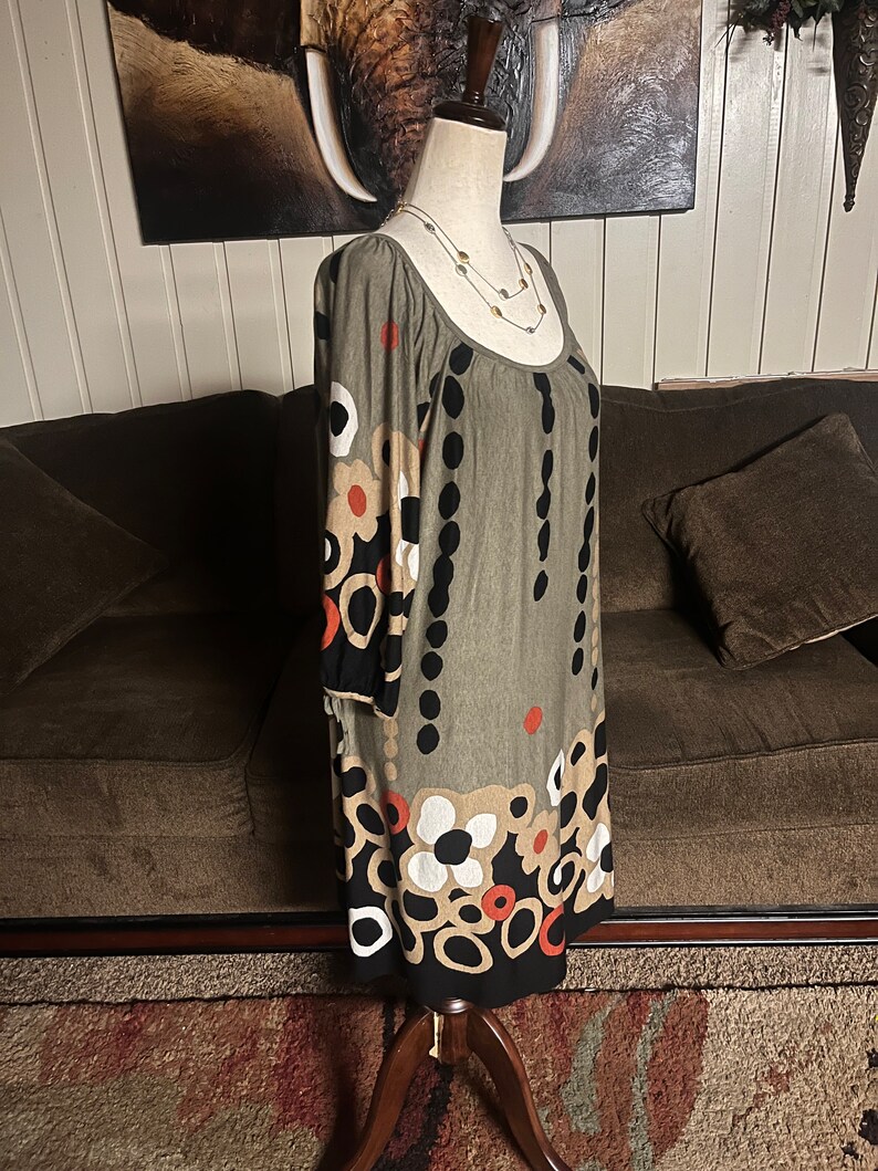 May include: A green, black, white, and orange patterned dress with a scoop neck and 3/4 sleeves. The dress is on a mannequin and is displayed against a brown couch.