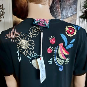 Puede incluir: Una camiseta negra de manga corta con bordados coloridos en la parte posterior. El dise&ntilde;o incluye un girasol amarillo, fresas rojas, un pl&aacute;tano y varios patrones florales y abstractos. Una etiqueta blanca cuelga del cuello.