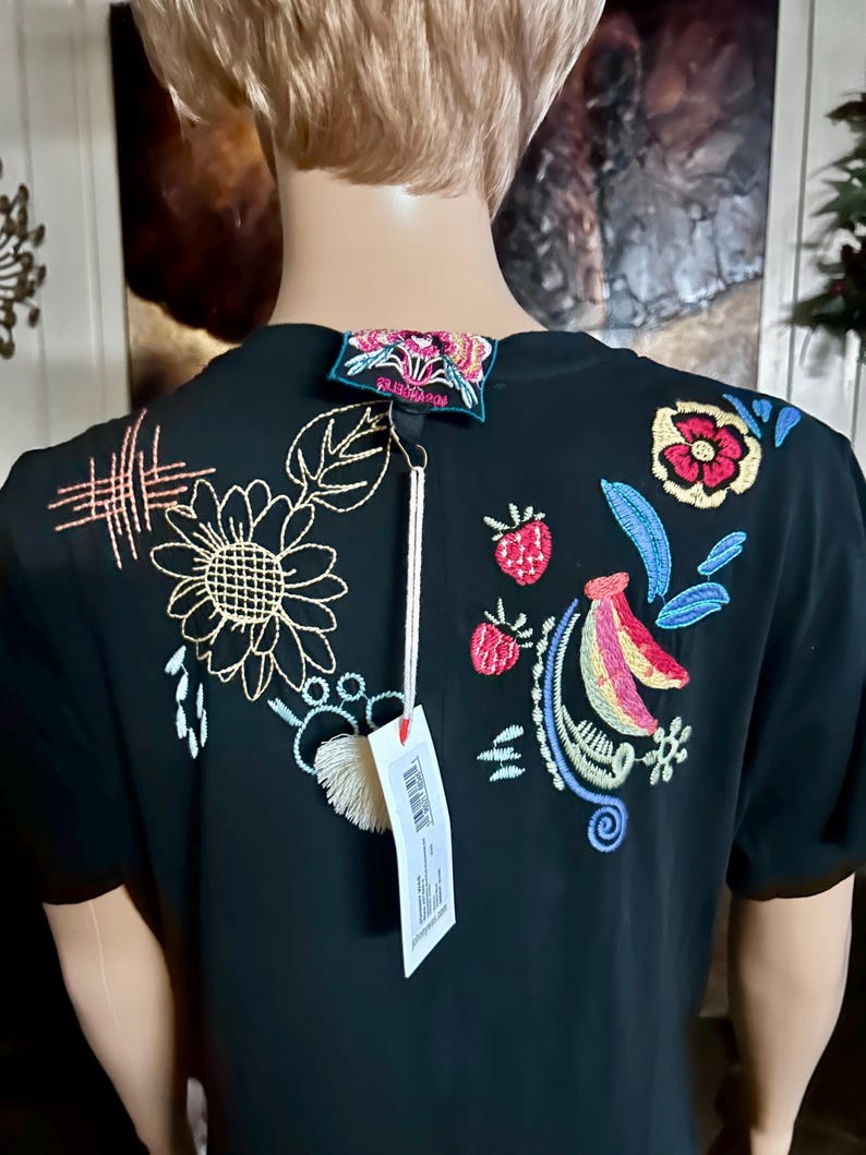 Puede incluir: Una camiseta negra de manga corta con bordados coloridos en la parte posterior. El dise&ntilde;o incluye un girasol amarillo, fresas rojas, un pl&aacute;tano y varios patrones florales y abstractos. Una etiqueta blanca cuelga del cuello.