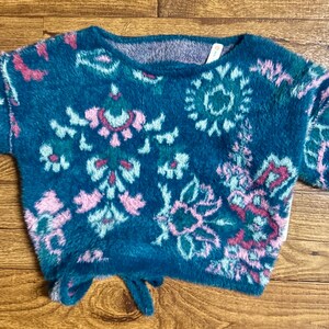 May include: A teal blue fuzzy sweater with a pink, green, and white floral pattern. The sweater has a tie at the bottom.