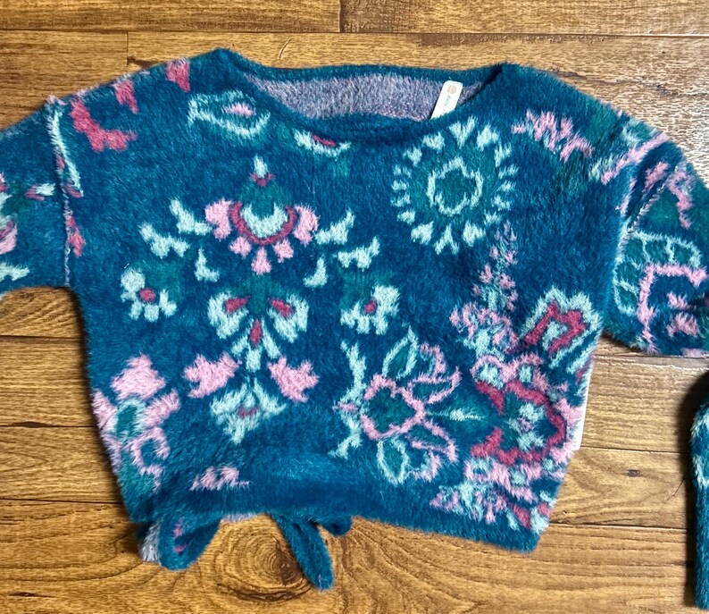 May include: A teal blue fuzzy sweater with a pink, green, and white floral pattern. The sweater has a tie at the bottom.