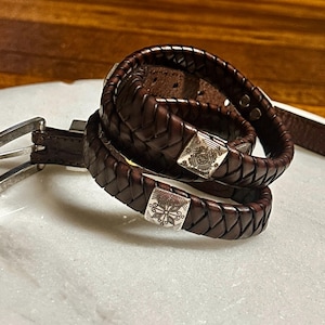 K&ouml;nnte beinhalten: Ein dunkelbraunes Lederg&uuml;rtel- und passendes geflochtenes Armband-Set. Der G&uuml;rtel hat eine silberfarbene Schnalle. Das Armband hat quadratische silberfarbene Verschl&uuml;sse mit dekorativen Designs. Das Leder hat ein gewebtes Muster.