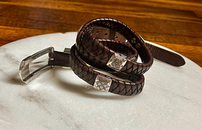 K&ouml;nnte beinhalten: Ein dunkelbraunes Lederg&uuml;rtel- und passendes geflochtenes Armband-Set. Der G&uuml;rtel hat eine silberfarbene Schnalle. Das Armband hat quadratische silberfarbene Verschl&uuml;sse mit dekorativen Designs. Das Leder hat ein gewebtes Muster.