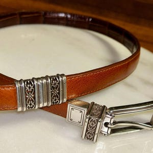 May include: A brown leather belt with a silver buckle and decorative accents. The belt has a rectangular buckle with an ornate design and a matching silver loop. The leather is a medium brown color.