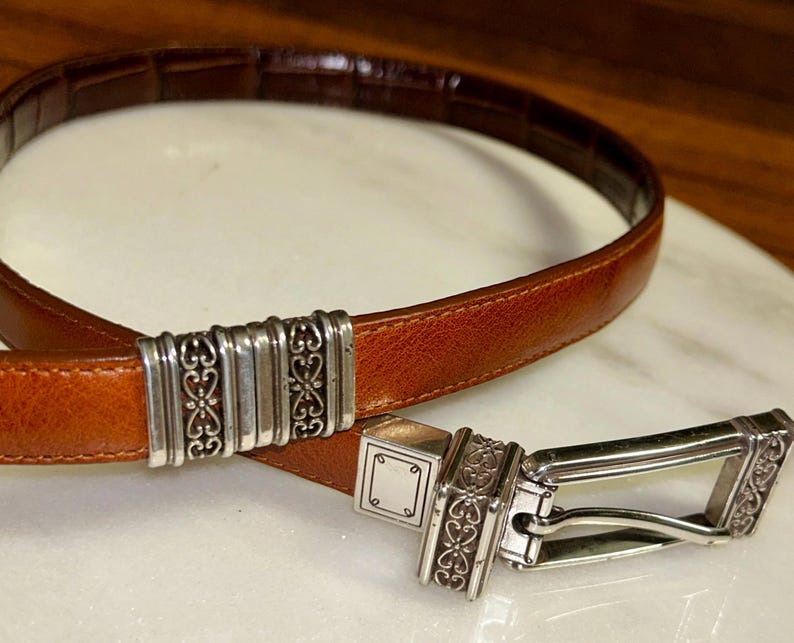 May include: A brown leather belt with a silver buckle and decorative accents. The belt has a rectangular buckle with an ornate design and a matching silver loop. The leather is a medium brown color.