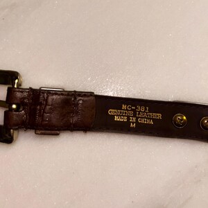 May include: A brown leather belt with a gold-colored buckle and hardware. The belt has a textured surface and is labeled "MC-381 GENUINE LEATHER MADE IN CHINA M". The belt is medium size.