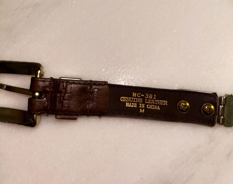 May include: A brown leather belt with a gold-colored buckle and hardware. The belt has a textured surface and is labeled "MC-381 GENUINE LEATHER MADE IN CHINA M". The belt is medium size.