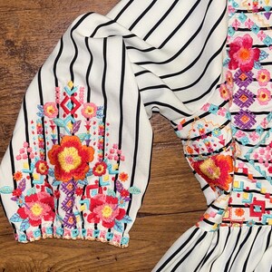 May include: White and black striped dress with colourful embroidered flowers and geometric patterns. The dress has a ruffled hem.