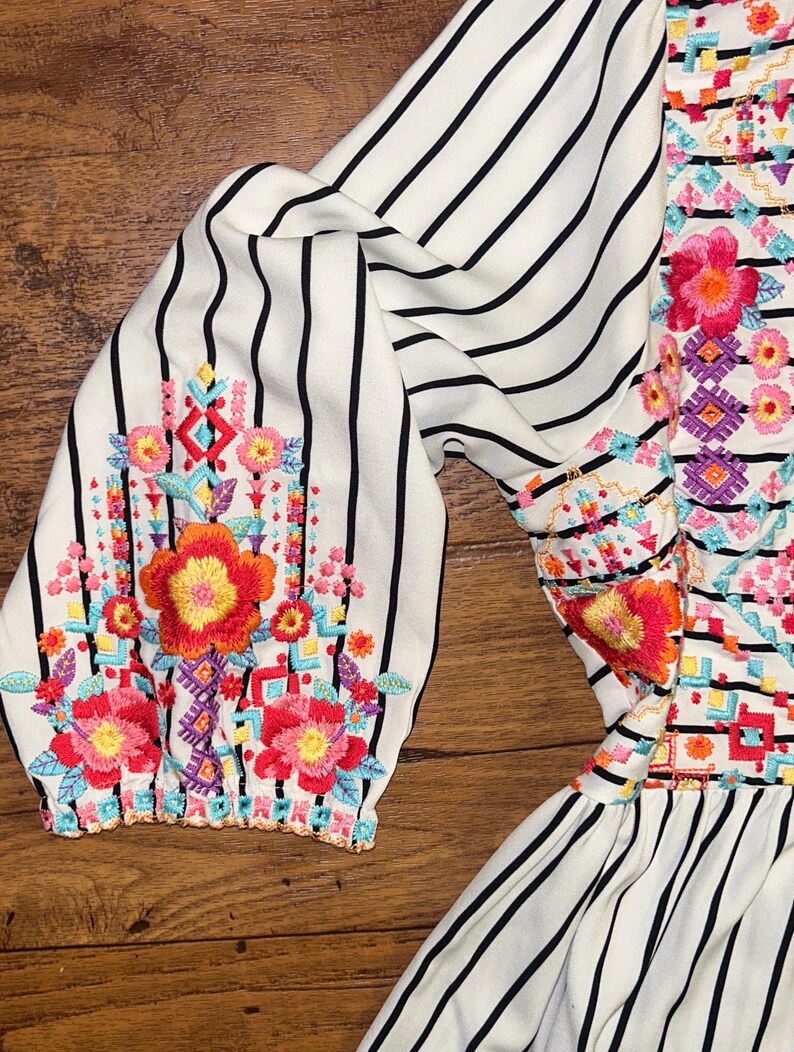 May include: White and black striped dress with colourful embroidered flowers and geometric patterns. The dress has a ruffled hem.