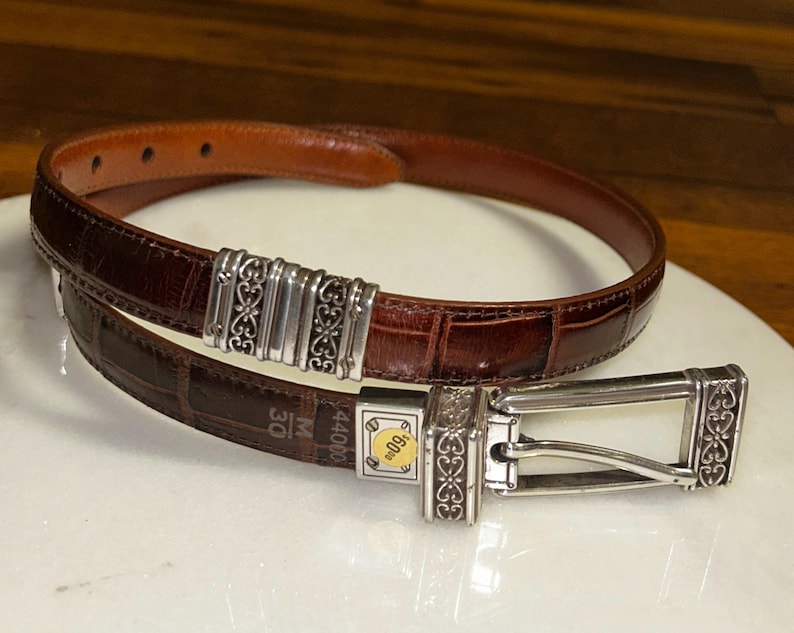 May include: A brown leather belt with a crocodile texture. The belt has a silver buckle and decorative silver accents. The belt is approximately 30 inches in length.