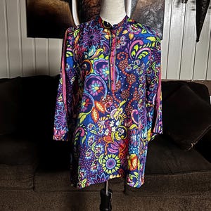 May include: A long-sleeved tunic with a vibrant floral pattern in shades of blue, purple, pink, yellow, and orange. The tunic has a mandarin collar and a button-down front. The sleeves are long and the fabric appears to be a lightweight material.