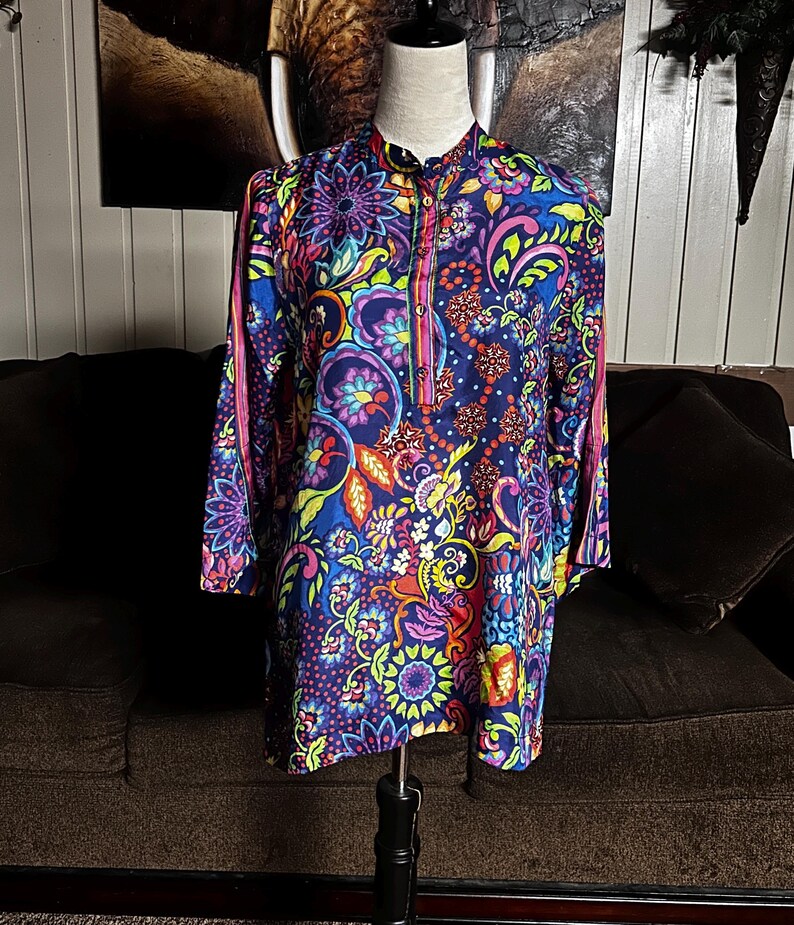 May include: A long-sleeved tunic with a vibrant floral pattern in shades of blue, purple, pink, yellow, and orange. The tunic has a mandarin collar and a button-down front. The sleeves are long and the fabric appears to be a lightweight material.