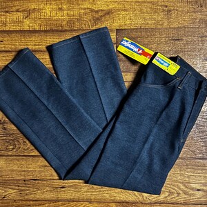 Może przedstawiać: Ciemnoniebieskie dżinsy Wrangler z denimu o klasycznym kroju, z prostą nogawką oraz zapięciem na guzik i zamek błyskawiczny. Dżinsy mają ż&oacute;łto-niebieską metkę Wrangler. Dżinsy leżą płasko na drewnianej powierzchni.