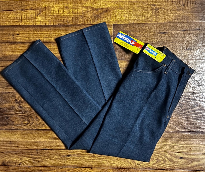 Może przedstawiać: Ciemnoniebieskie dżinsy Wrangler z denimu o klasycznym kroju, z prostą nogawką oraz zapięciem na guzik i zamek błyskawiczny. Dżinsy mają ż&oacute;łto-niebieską metkę Wrangler. Dżinsy leżą płasko na drewnianej powierzchni.