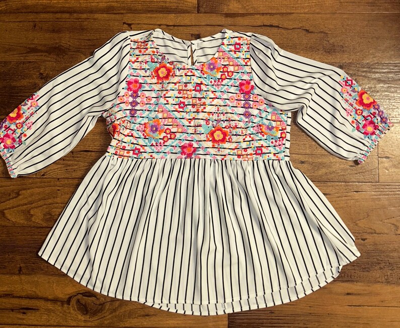 May include: White and black striped blouse with a floral embroidered yoke. The embroidery features pink, red, blue, and yellow flowers.