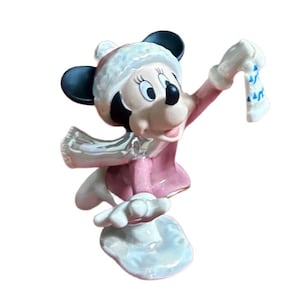 Może przedstawiać: Ceramiczna figurka Myszki Minnie w zimowym stroju. Ma na sobie r&oacute;żowy płaszcz, czapkę i szalik, z biało-niebieską rękawicą. Figurka ma czarne uszy i uśmiechniętą twarz, stojąc na śnieżnobiałej podstawie.