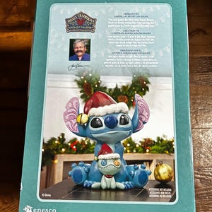 May include: A blue Disney Traditions figurine of Stitch wearing a Santa hat, holding a small doll, and sitting on a base. The figurine is on a box with text that reads "Designed by American Artist Jim Shore" and "Disney Traditions".