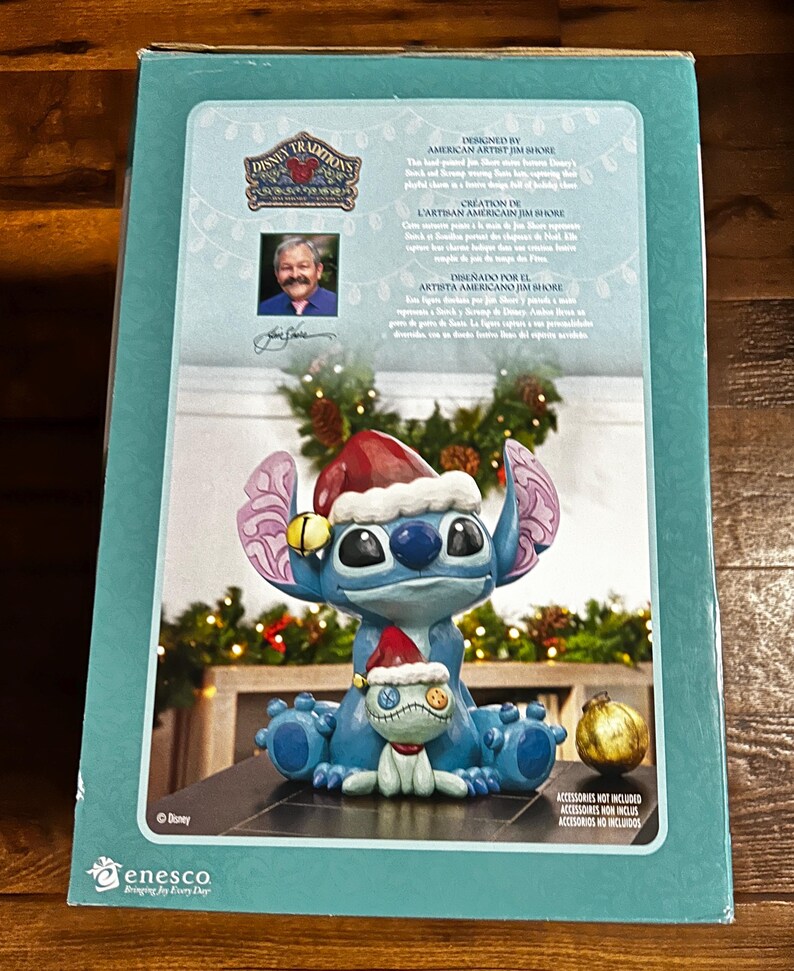 May include: A blue Disney Traditions figurine of Stitch wearing a Santa hat, holding a small doll, and sitting on a base. The figurine is on a box with text that reads "Designed by American Artist Jim Shore" and "Disney Traditions".