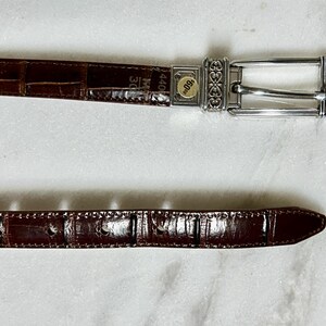 May include: A brown leather belt with a silver buckle. The belt has a crocodile texture and is marked with the size 30. The buckle has an ornate design. The belt is approximately 44 inches long.