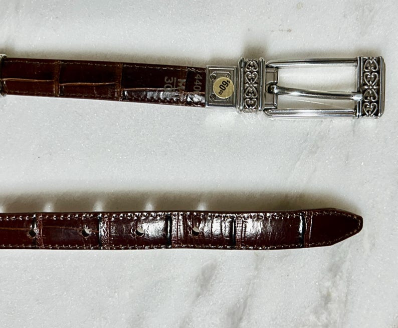 May include: A brown leather belt with a silver buckle. The belt has a crocodile texture and is marked with the size 30. The buckle has an ornate design. The belt is approximately 44 inches long.