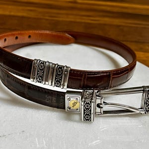 May include: A brown leather belt with a silver buckle and decorative accents. The belt has a crocodile texture and a silver buckle with a gold accent. The belt is approximately 1.25 inches wide.