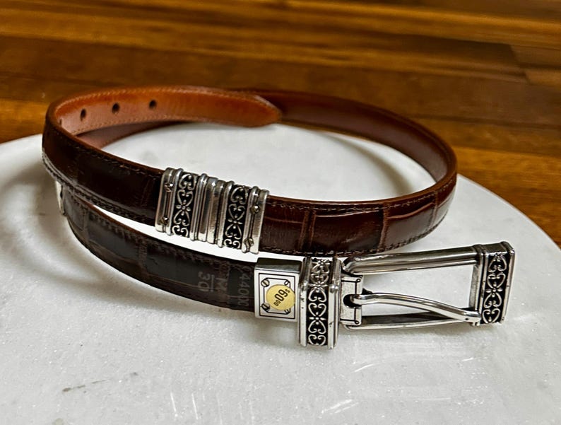 May include: A brown leather belt with a silver buckle and decorative accents. The belt has a crocodile texture and a silver buckle with a gold accent. The belt is approximately 1.25 inches wide.