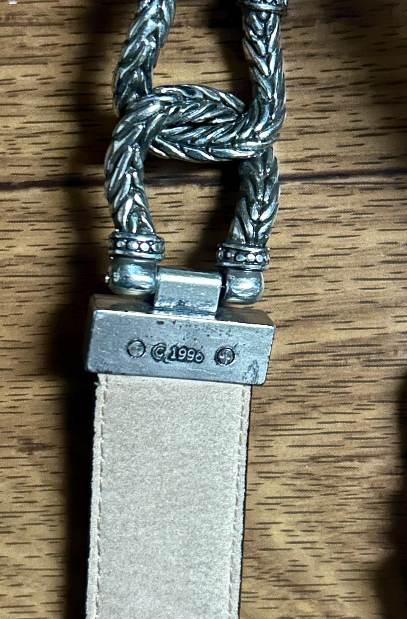 Puede incluir: Una hebilla de cintur&oacute;n de plata con un dise&ntilde;o trenzado y el texto "&copy;1996". La hebilla est&aacute; unida a un cintur&oacute;n de cuero beige.