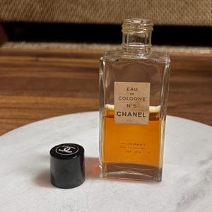 May include: A rectangular glass bottle of Chanel No. 5 Eau de Cologne with a black cap. The bottle has a beige label with black text. The cologne inside is amber-colored. The black cap has a white Chanel logo.