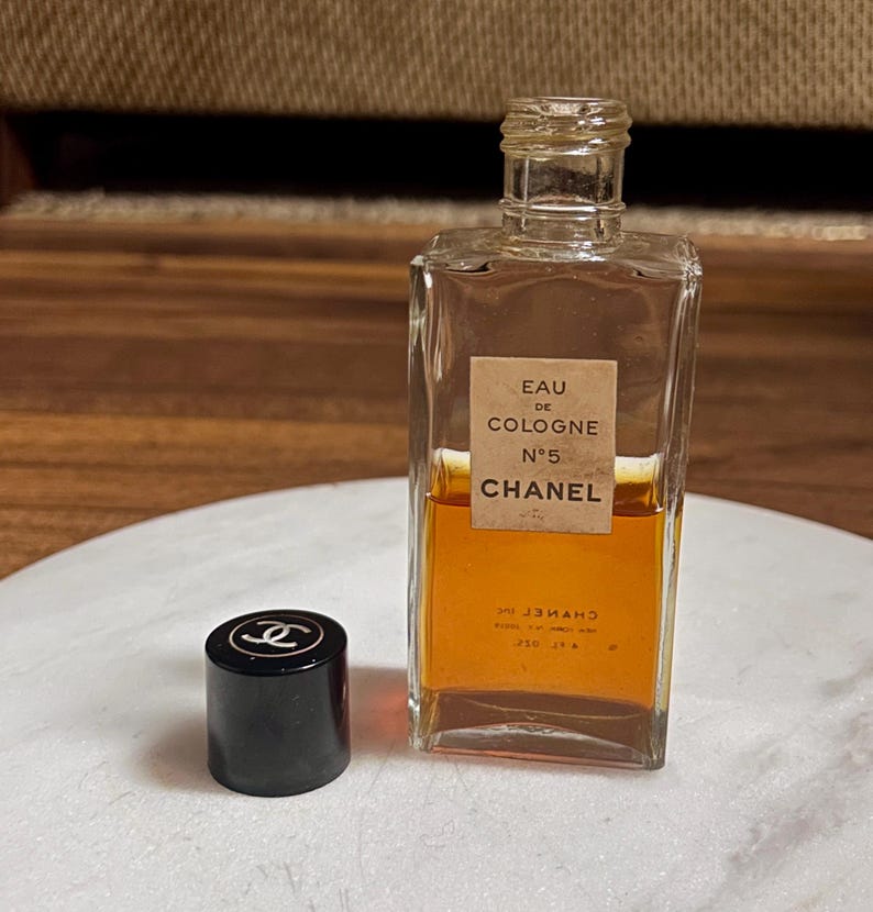 May include: A rectangular glass bottle of Chanel No. 5 Eau de Cologne with a black cap. The bottle has a beige label with black text. The cologne inside is amber-colored. The black cap has a white Chanel logo.