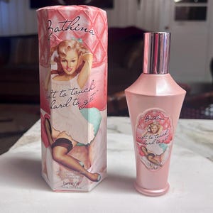 May include: A pink Bathina perfume bottle and matching box. The box features a vintage pin-up illustration with the text "soft to touch, hard to get." The bottle has a pink cap and a label with the same illustration.