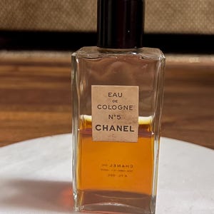 May include: A rectangular glass bottle of Chanel No. 5 Eau de Cologne. The bottle has a dark brown cap and a label with the text "EAU DE COLOGNE N&deg;5 CHANEL". The liquid inside is amber-colored.