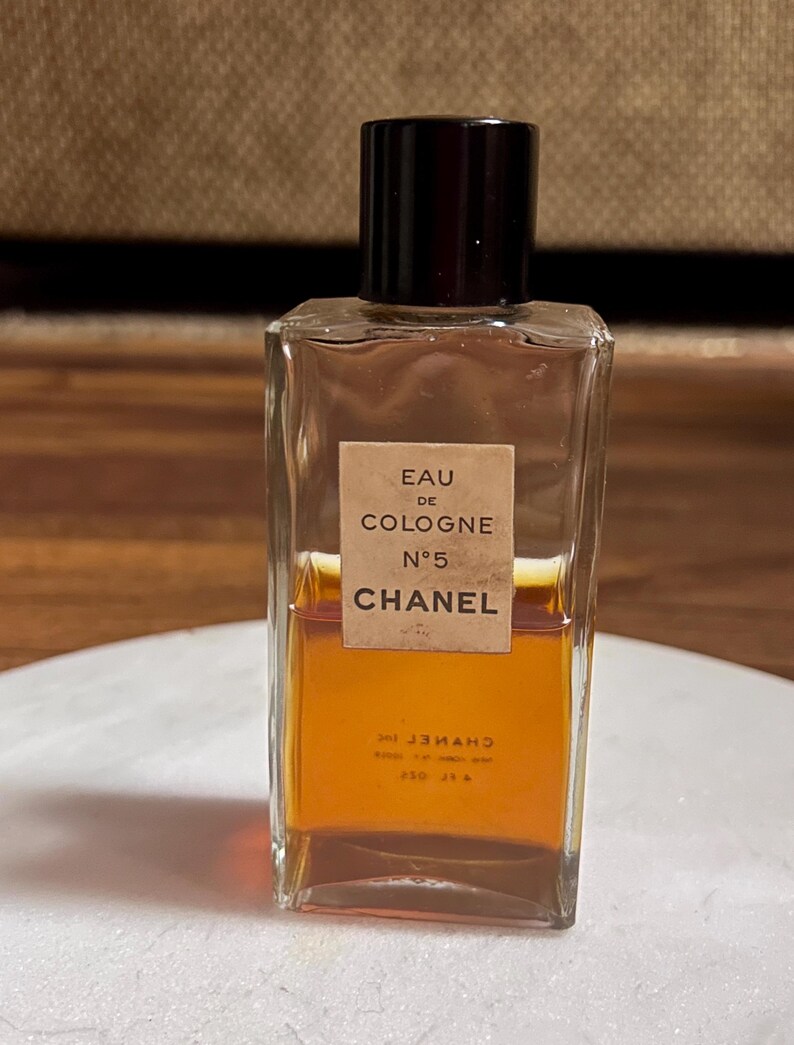 May include: A rectangular glass bottle of Chanel No. 5 Eau de Cologne. The bottle has a dark brown cap and a label with the text "EAU DE COLOGNE N&deg;5 CHANEL". The liquid inside is amber-colored.