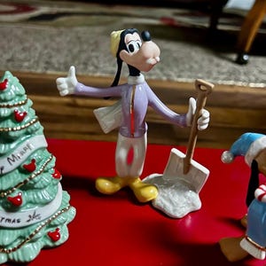 Może przedstawiać: Figurka Goofy'ego w zimowym stroju, trzymająca łopatę, z gestem kciuka w g&oacute;rę. Figurka ma na sobie fioletową kurtkę, białe spodnie i ż&oacute;łte buty. W tle choinka i kolejna figurka.