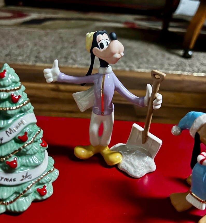 Może przedstawiać: Figurka Goofy'ego w zimowym stroju, trzymająca łopatę, z gestem kciuka w g&oacute;rę. Figurka ma na sobie fioletową kurtkę, białe spodnie i ż&oacute;łte buty. W tle choinka i kolejna figurka.