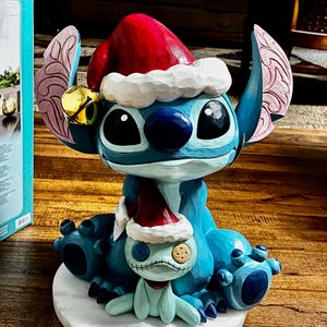 May include: A blue Stitch figurine wearing a red Santa hat with white trim and a gold bell. A smaller, light blue creature with a stitched face and a Santa hat sits in front of Stitch. The figurine is on a white base.
