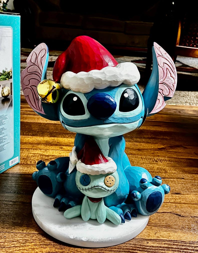 May include: A blue Stitch figurine wearing a red Santa hat with white trim and a gold bell. A smaller, light blue creature with a stitched face and a Santa hat sits in front of Stitch. The figurine is on a white base.